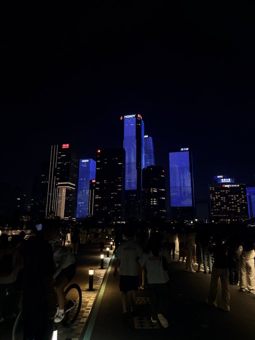 A nighttime cityscape of illuminated skyscrapers including 'HENGRUI' and 'CR GROUP', with crowds of people gathered along a waterfront promenade.