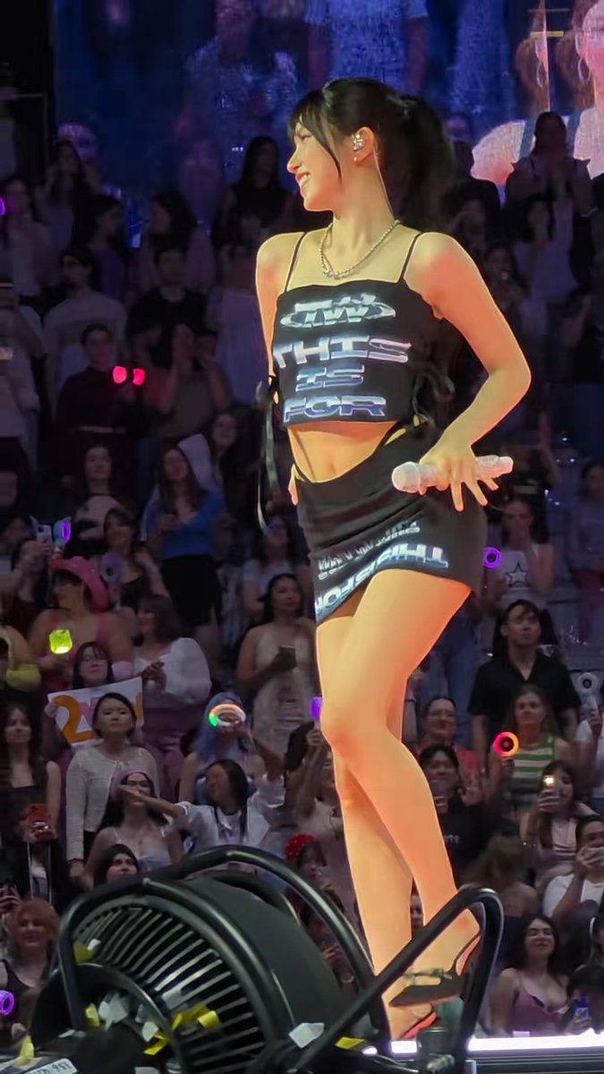 A performer on stage wearing a black crop top reading 'THIS IS FOR' and a matching skirt, holding a microphone in front of a cheering crowd holding light sticks.