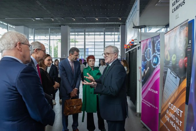 The image shows a group of people gathered around a booth at an event, possibly a conference or exhibition. The booth features a poster for the #IOTA roller banners project supported by the Imperial College, which was presented by researchers to Lord Vallance and Marc Lemaître. There are several people in the scene, including one man wearing a suit who is talking to another person standing next to him. A handbag can also be seen on the ground near the booth. The image suggests that the attendees of this event might be interested in the #IOTA project or its implications for various fields such as health and technology.