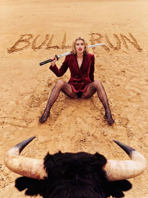 A blonde woman in a red velvet blazer and fishnet stockings poses with a katana in a sandy arena, with 'BULL RUN' written in the sand behind her and a bull's horns visible in the foreground.
