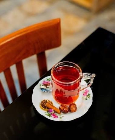 A glass cup of red tea sitting on a floral-patterned saucer with dried fruits and a cinnamon stick, placed on a black table next to a wooden chair.