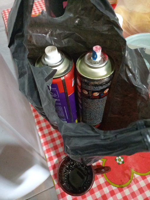 Two aerosol spray cans sit inside a black plastic bag on a red and white checkered tablecloth next to a dark mug.