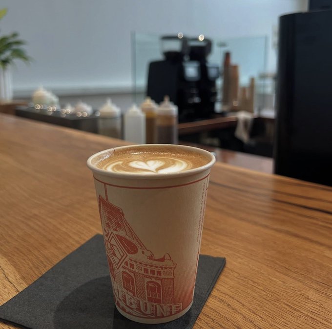 A close-up photograph of a takeaway coffee cup with latte art, sitting on a black coaster on a wooden cafe counter; the cup features a red architectural illustration and the partial text "BUNE".