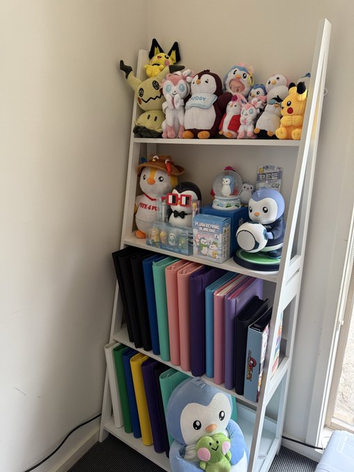 A white ladder-style shelf holds plush toys, figurines, boxed collectibles, and rows of brightly colored binders.