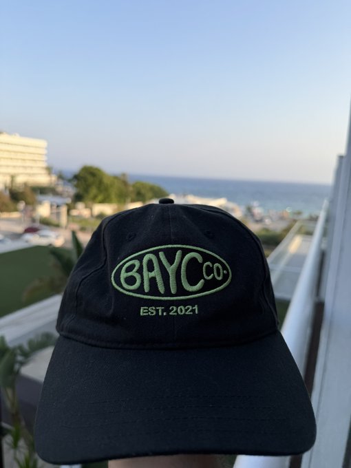 A black baseball cap with green embroidered text 'BAYC CO. EST. 2021' is photographed outdoors with a coastal view and building visible in the background.