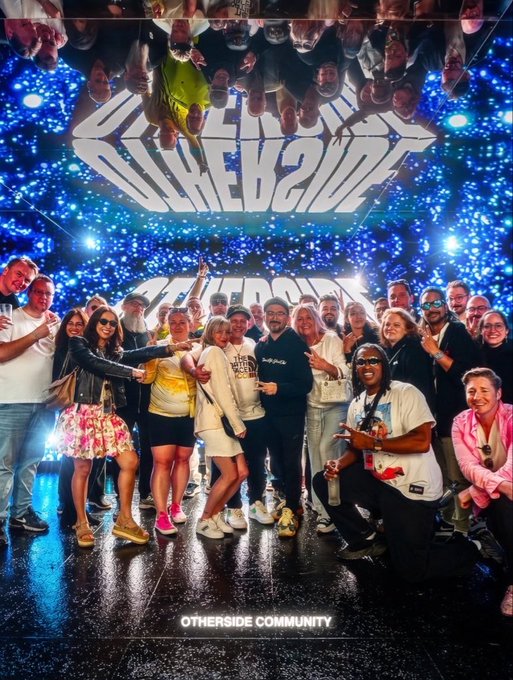 A group photo of people posing under illuminated 'OTHERSIDE' signage in a starry blue mirrored room, with 'OTHERSIDE COMMUNITY' written at the bottom.