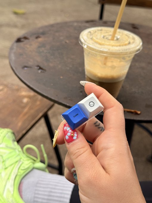 A hand with red and white nail art holds a blue and white Cube keychain in front of an iced coffee on a small round table.
