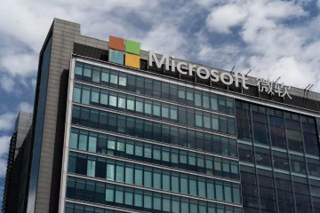 A modern glass office building with the Microsoft logo (four colored squares) and 'Microsoft 微软' signage on its facade under a partly cloudy sky.