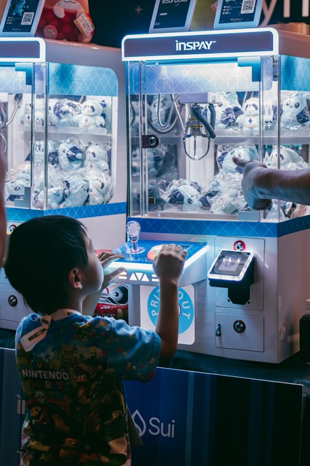 A young boy in a Nintendo-themed shirt plays with a claw machine labeled 'inSPay' containing plush toys, with the 'Sui' blockchain logo visible on the front.