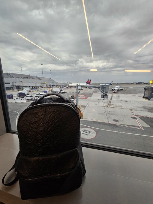 A black textured backpack sits on a windowsill at an airport terminal, looking out over the tarmac where planes and ground vehicles are visible under a cloudy sky.