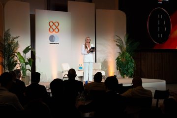 A woman speaking on a stage at a corporate event with a logo backdrop, holding a microphone while addressing an audience.