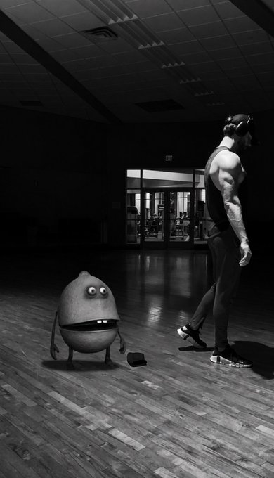 A black and white image of a muscular man with headphones walking on a gym floor past a cartoonish, animated egg-shaped character with big eyes.