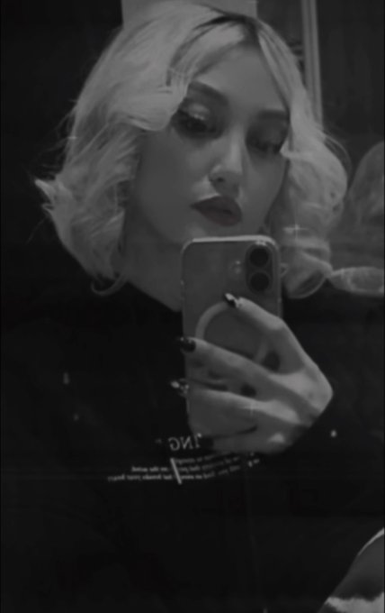 A black and white filtered selfie of a blonde woman taking a mirror photo with her smartphone, showing dark nail polish and makeup.