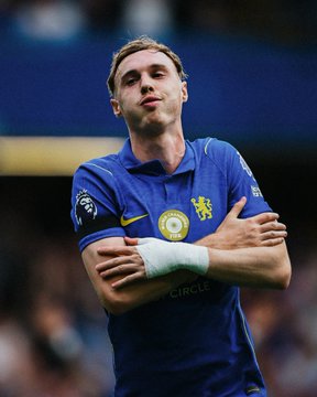 A Chelsea player in the blue jersey celebrates a goal with his arms crossed in his signature celebration pose, fitting the post's theme of celebrating a winning sports betting score prediction. The jersey displays "CIRCLE" as a sponsor and features the Chelsea club crest.