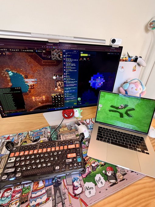 A desk setup featuring a large monitor running a game or virtual environment alongside a chat window, with a MacBook displaying a green game screen, a colorful mechanical keyboard, and a gaming mouse on a patterned mousepad.
