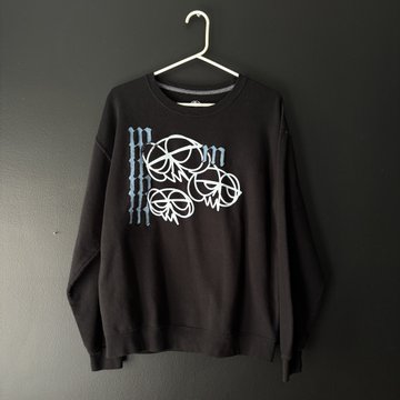 A black crewneck sweatshirt hanging on a white plastic hanger against a dark gray wall, featuring a white graphic design of three stylized faces.