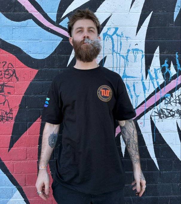 A bearded man wearing a black "TLOT" t-shirt exhales a cloud of smoke while standing in front of a brick wall covered in colorful blue, pink, and black graffiti.