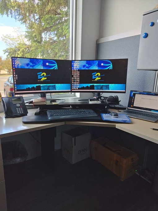 An office desk setup features dual monitors displaying an 'E2' logo, a LG laptop, a keyboard, mouse, and telephone, with a large window providing natural light.