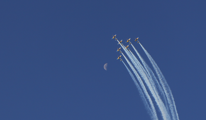 A formation of jets flies diagonally across a deep blue sky, leaving white contrails, with a crescent moon visible in the background.