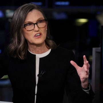 A woman with shoulder-length brown hair and glasses, wearing a black jacket with white trim, gesturing with her hand while speaking.