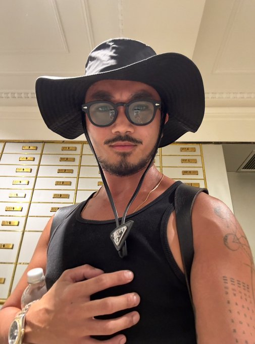 A stylish man wearing a wide-brim black Prada hat, sunglasses, and a black tank top takes a selfie, accessorized with a Prada necklace and gold watch, while holding a water bottle in a room lined with safe deposit boxes. The image contrasts the post's crypto industry milestones—growing the $STX community and developer ecosystem at Stacks—with a polished, fashionable personal aesthetic.