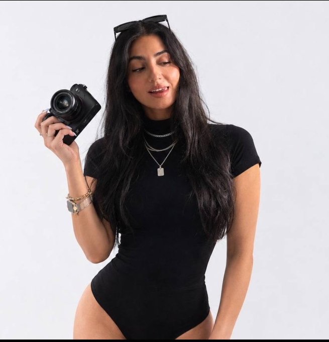 A young woman with long dark hair wearing a black bodysuit holds a camera near her face, with sunglasses perched on top of her head and a silver padlock necklace around her neck.