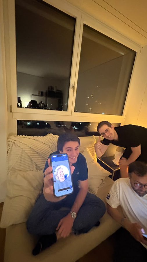 Three men in a living room at night; one seated on a couch holds up a phone displaying a video call or profile picture, while another leans over the back of the couch smiling.