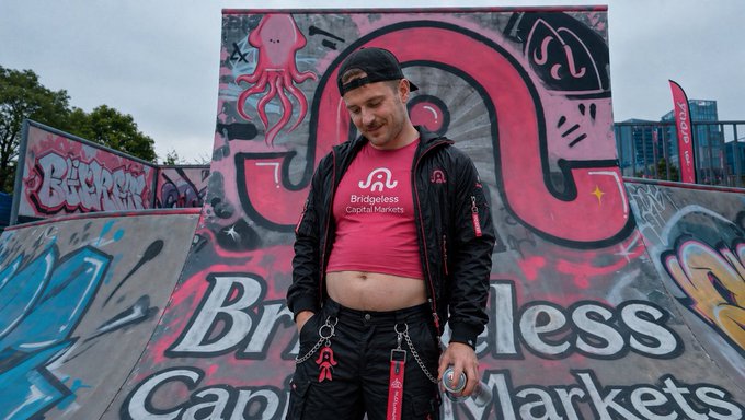 A man wearing a black cap and jacket over a pink 'Bridgeless Capital Markets' t-shirt stands in front of a colorful graffiti-covered skate ramp.