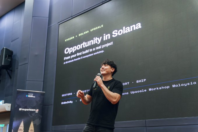 A young man with curly hair stands on stage holding a microphone, smiling while presenting. Behind him, a large screen displays the title 'Opportunity in Solana - From your first build to a real project' for the Kyzen × Solana Upscale Workshop Malaysia.