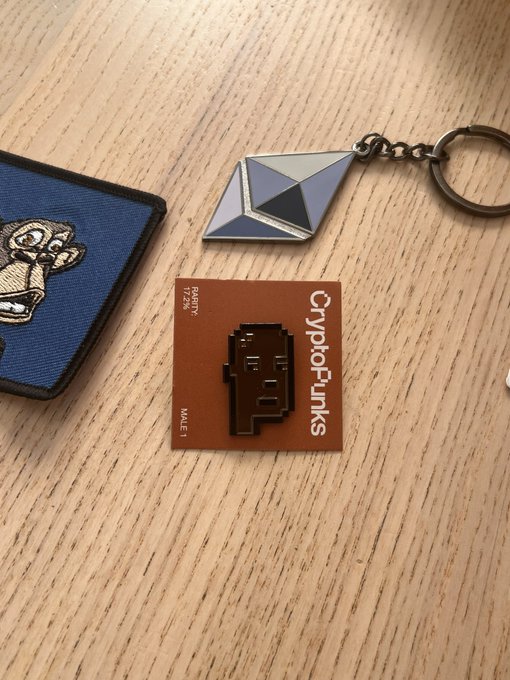A CryptoPunks enamel pin and keychain displayed on a wooden surface, featuring the iconic pixel-art 'Male 1' character on its original '1 of 24 Rarity' backing card.
