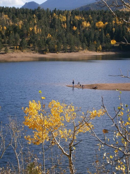 Two small figures stand on a narrow strip of land jutting into a calm blue lake, framed by golden aspen branches in the foreground and a forested mountain backdrop in autumn colors. The serene scene captures the quiet reflection and fresh-start energy of stepping into a new month, with the expansive waters and fall foliage echoing the hopeful tone of embracing the months ahead.