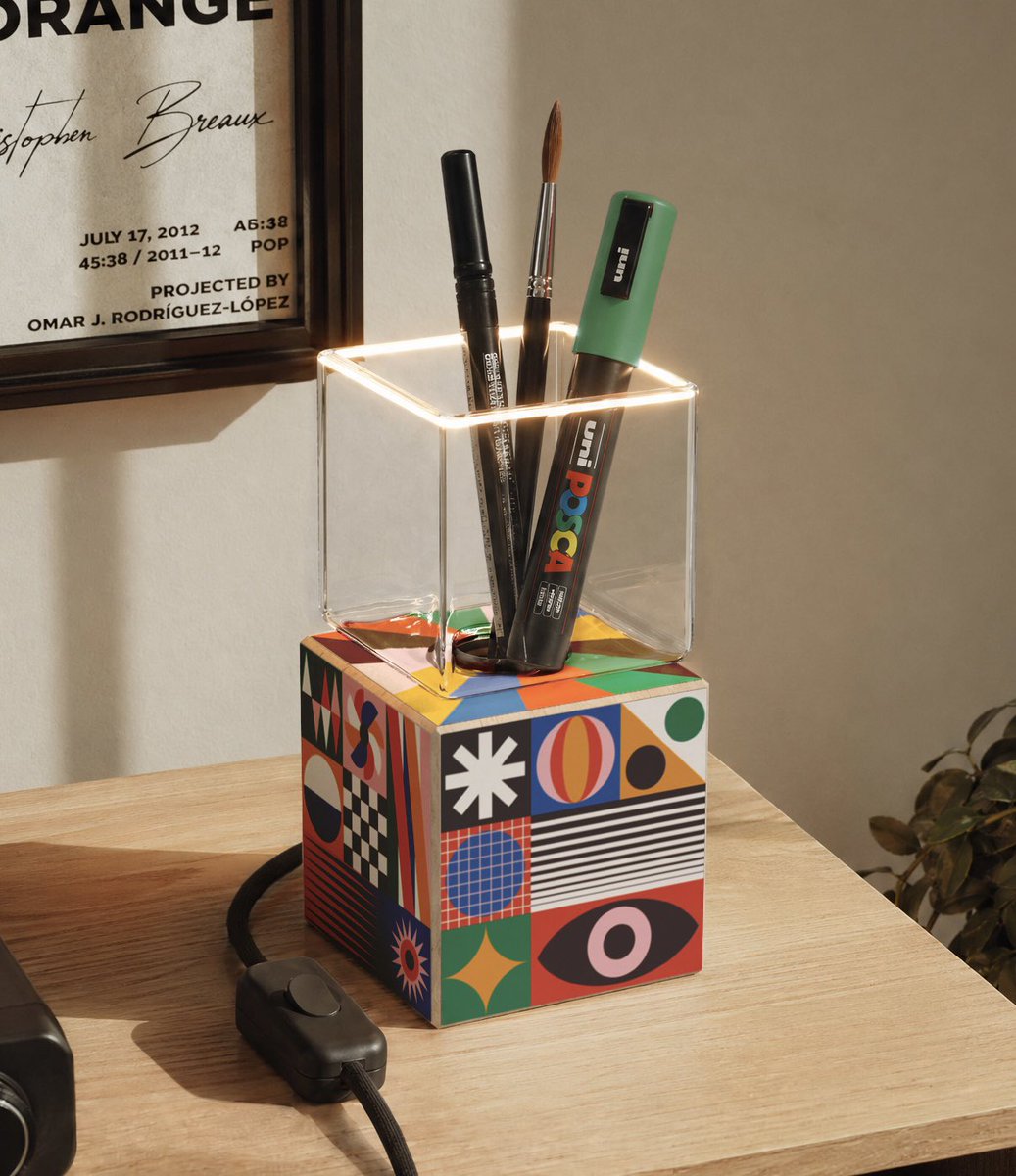 A desk scene features a clear glass pen holder sitting atop a colorful cube decorated with geometric shapes, containing POSCA paint markers and a Uni marker. A framed poster on the wall behind reads 'Stephen Breaux', 'JULY 17, 2012 A6:38', '45:38 / 2011–12 POP', 'PROJECTED BY OMAR J. RODRÍGUEZ-LÓPEZ'.