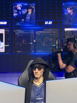 A trader wearing a "roblox" jersey, sunglasses, and headphones sits at a multi-monitor trading station displaying a "03.32" timer and live market charts, while another trader films him with a phone—capturing the lively atmosphere of the IRL trading competition mentioned in the post.