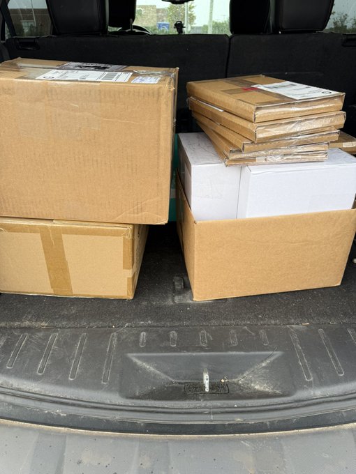 The trunk of a vehicle is loaded with cardboard boxes and padded manila envelopes, presumably containing VeeFriends comic books ready for shipping worldwide. A shipping label is partially visible on the top box, tying into the post about fans who purchased comics from the midnight release of Comic 19.