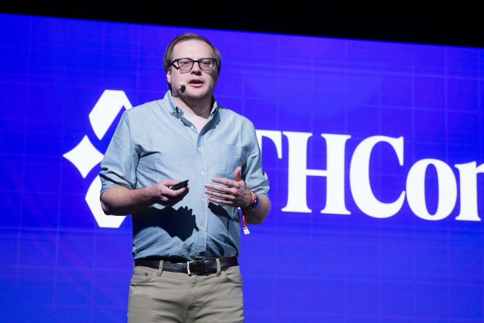The image features a man standing on stage, wearing glasses and a blue shirt with khaki pants. He is holding a microphone and appears to be giving a speech or presentation. The background of the image shows a large screen displaying the words "THCHCON" in white letters against a blue backdrop. The text suggests that the speaker might be discussing the benefits of using blockchain technology, specifically for financial institutions.