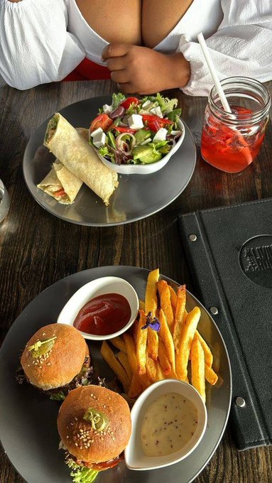 An overhead photo of a restaurant meal featuring a plate with two small burgers, sweet potato fries, and two dipping sauces, alongside another plate with a wrap, salad, and a mason jar of iced red drink.