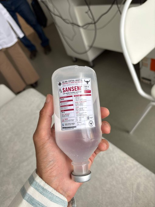 A hand holds an IV fluid bag labeled 'R. SANSEM' and 'SOLAPA CENTRAL HOSPITAL' in what appears to be a clinical setting.