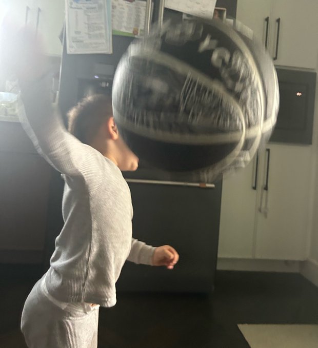 A motion-blurred photo of a person in a grey long-sleeve shirt holding or throwing a basketball in a kitchen setting.