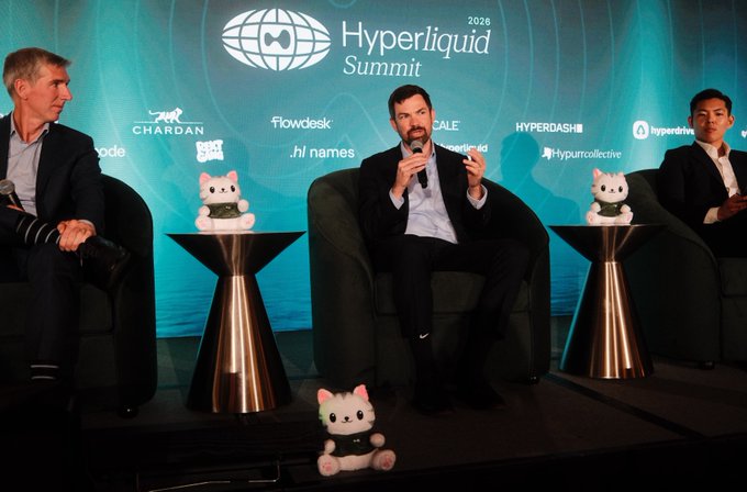 Three panelists sit in green armchairs on stage at the "Hyperliquid Summit 2026," with the central speaker holding a microphone and gesturing, while mascot plush figures are displayed on side tables and at the base of the stage. The backdrop features sponsor logos including Chardan, Flowdesk, Scale, HYPERDASH, Hyperdrive, .htf names, and Hyperuncollective, visually tying to the caption's announcement of major financial firms like a16zcrypto, Goldman Sachs, Bloomberg, JPMorgan, and Multicoin attending the event.