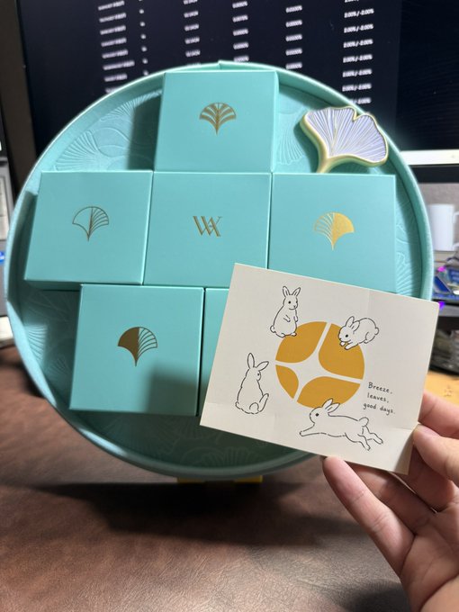 A hand holds a card featuring playful illustrations of rabbits and an orange with the text "Breezes, leaves, good days," while in the background a teal plate displays multiple small gift boxes adorned with ginkgo leaf motifs and a "W" monogram. The scene evokes the celebratory and gift-filled spirit of the AsterRonin program's six-month milestone, symbolizing appreciation for the 100 global ambassadors selected from over 1000+ applicants across 27 countries.