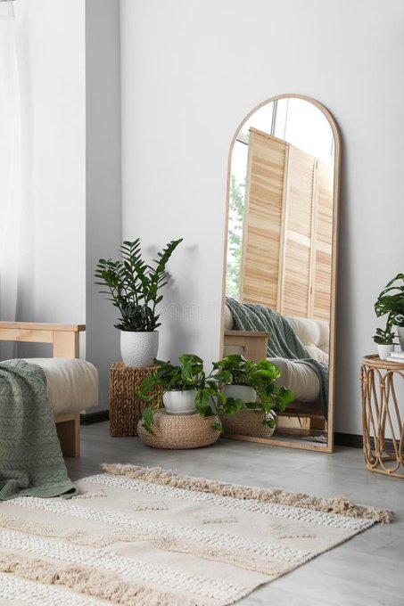 The image shows a living room featuring a large mirror hanging above a bed, surrounded by potted plants placed throughout the space. The room has white walls and a beige rug that adds warmth and comfort to the area. A chair can also be seen in the room, providing additional seating options for relaxation or conversation. The overall atmosphere of the room is inviting and cozy, with the mirror reflecting natural light from the window and the plants adding a touch of nature indoors.