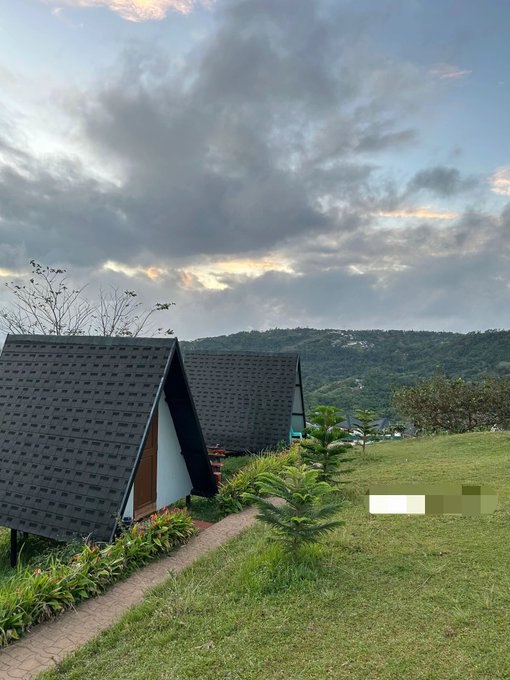 Scenic landscape view of A-frame cabins with dark roofs set on a grassy hillside, overlooking a mountainous region under a cloudy evening sky.
