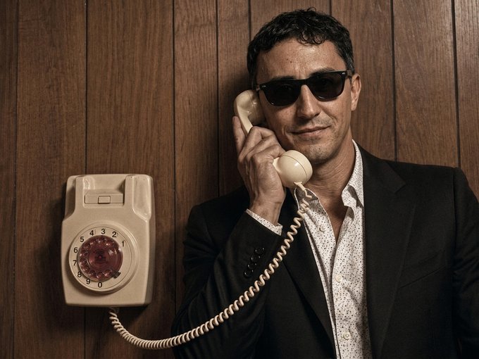 A man wearing sunglasses and a black suit jacket over a patterned shirt smiles while holding the receiver of a beige rotary phone mounted on a wood-paneled wall.