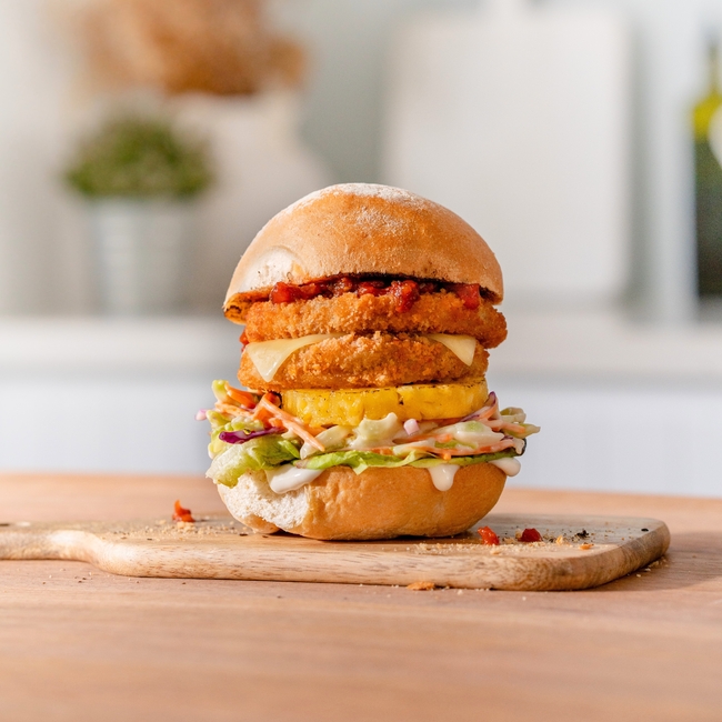 A tall chicken burger with a breaded chicken patty, cheese, a fried egg, coleslaw, and lettuce sits on a wooden serving board.