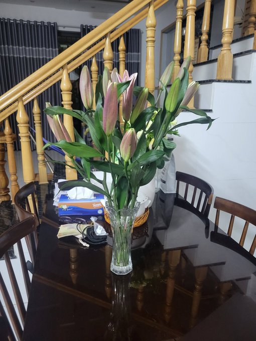 A vase of pink lilies sits on a glossy dark wooden dining table, with wooden stairs and chairs visible in the background. The image does not visually relate to the accompanying post about EthraShip's roadmap, as no text, charts, or project-related content appears in the scene.