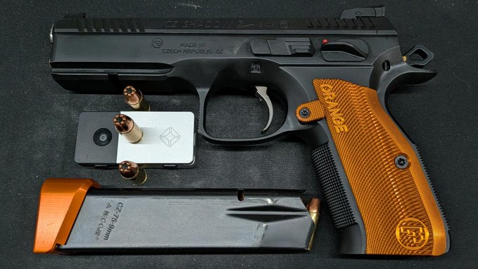 A black and orange CZ 75 Shadow 2 pistol is displayed with its magazine, three bullets, and a small tools box on a dark surface.