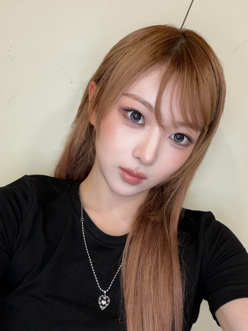 A young woman with long strawberry blonde hair, soft eye makeup, and a black t-shirt takes a selfie while wearing a silver heart pendant necklace. The image appears to be shared as a fromm post dated 260630, related to hashtags #AtHeart, #앳하트, #BOME, #봄, and #봄코어, suggesting the photo features a member of the group BOME (atHeart).