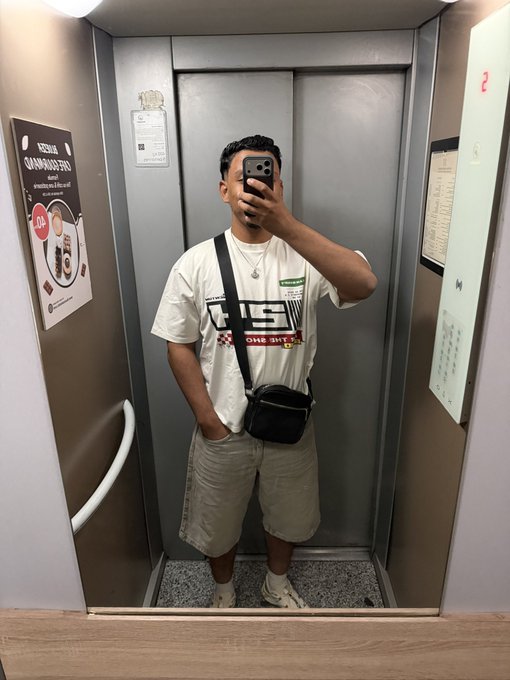 A man in a white graphic t-shirt and gray shorts takes a mirror selfie in an elevator, with his face obscured by his phone.