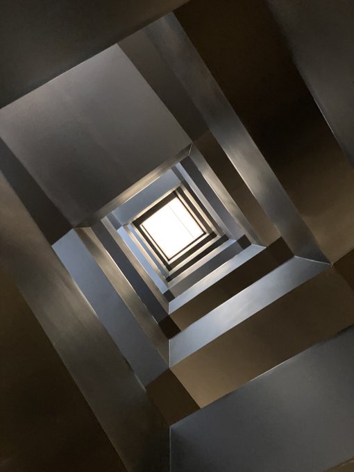 Abstract photograph looking straight up a square spiral staircase or architectural shaft toward a bright skylight opening at the top.