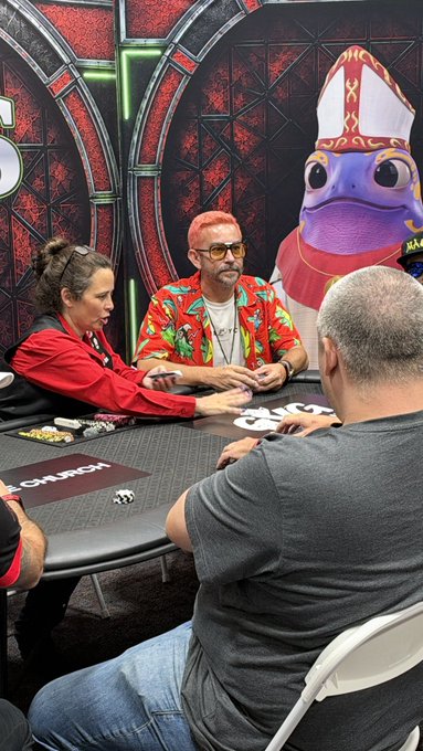 Four people play poker at a casino table with chips and cards visible, with a large purple duck mascot wearing a bishop's mitre in the background.